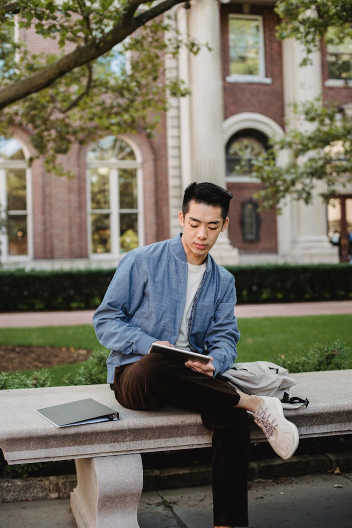 Focused Asian man browsing tablet while sitting on bench with backpack and folder in campus